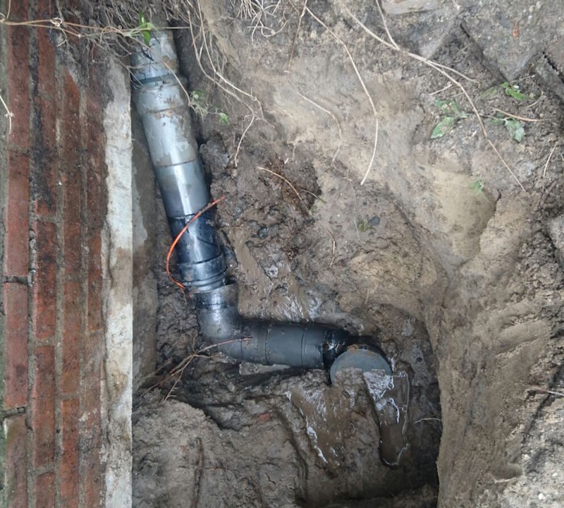 Op de foto zie je een opengegraven stuk grond naast een stenen gevel waarin een rioolbuis met verse lijmverbindingen en modderige resten is blootgelegd. Dit geeft een duidelijke indruk van hoe een leidingbreuk of verzakking in de afvoer gemakkelijk leidt tot ongewenste rioollucht in huis. De vochtige aarde rond de buis benadrukt het belang van tijdige lekdetectie om verdere schade aan fundament en vloeren te voorkomen. Door snel in te grijpen met professioneel herstel van het rioleringssysteem voorkom je niet alleen waterlekkage maar ook hardnekkige stankproblemen. Een grondige inspectie en reparatie vormen zo de sleutel tot een fris en droog binnenklimaat.